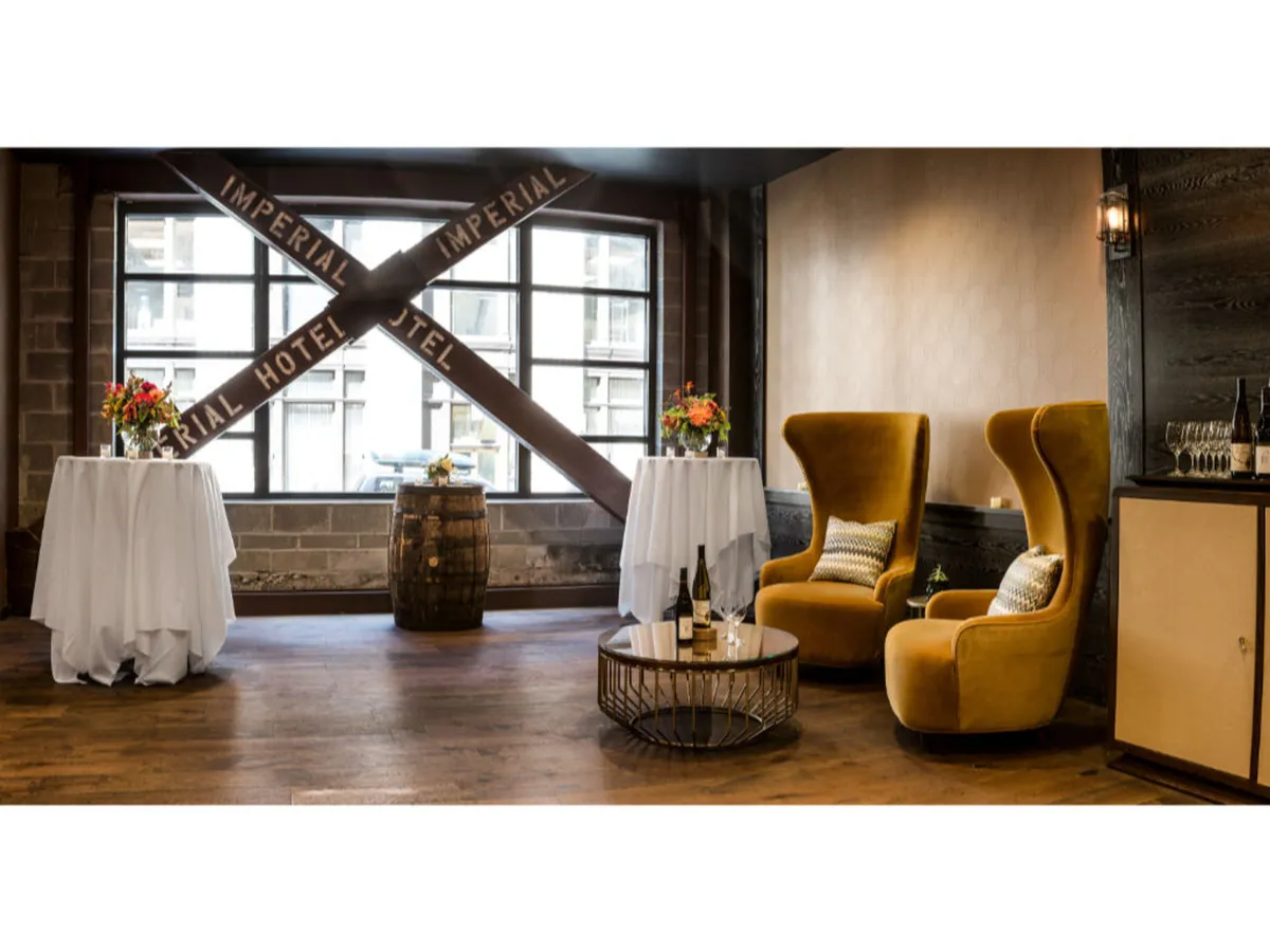 Inside, elegant yellow armchairs surround round tables adorned with fresh floral arrangements. A barrel accent table adds charm, and large windows with 'IMPERIAL HOTEL' signage flood the space with light. The setting is ideal for intimate gatherings, cocktail receptions, and relaxed social events.
