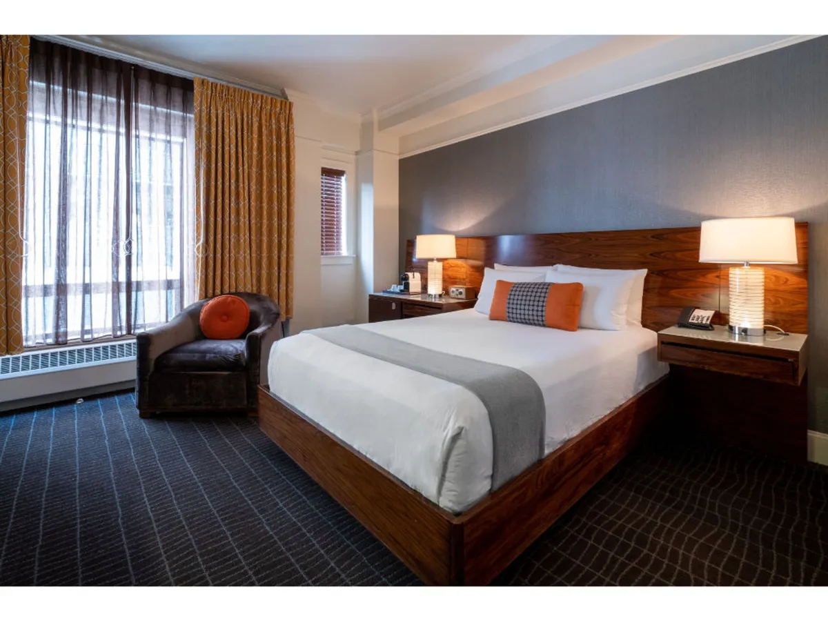The image showcases a stylish hotel room with a plush bed, soft natural light streaming through elegant curtains, a cozy armchair by the window, and a sleek nightstand—inviting spaces perfect for relaxation, reading, or enjoying a peaceful morning coffee.