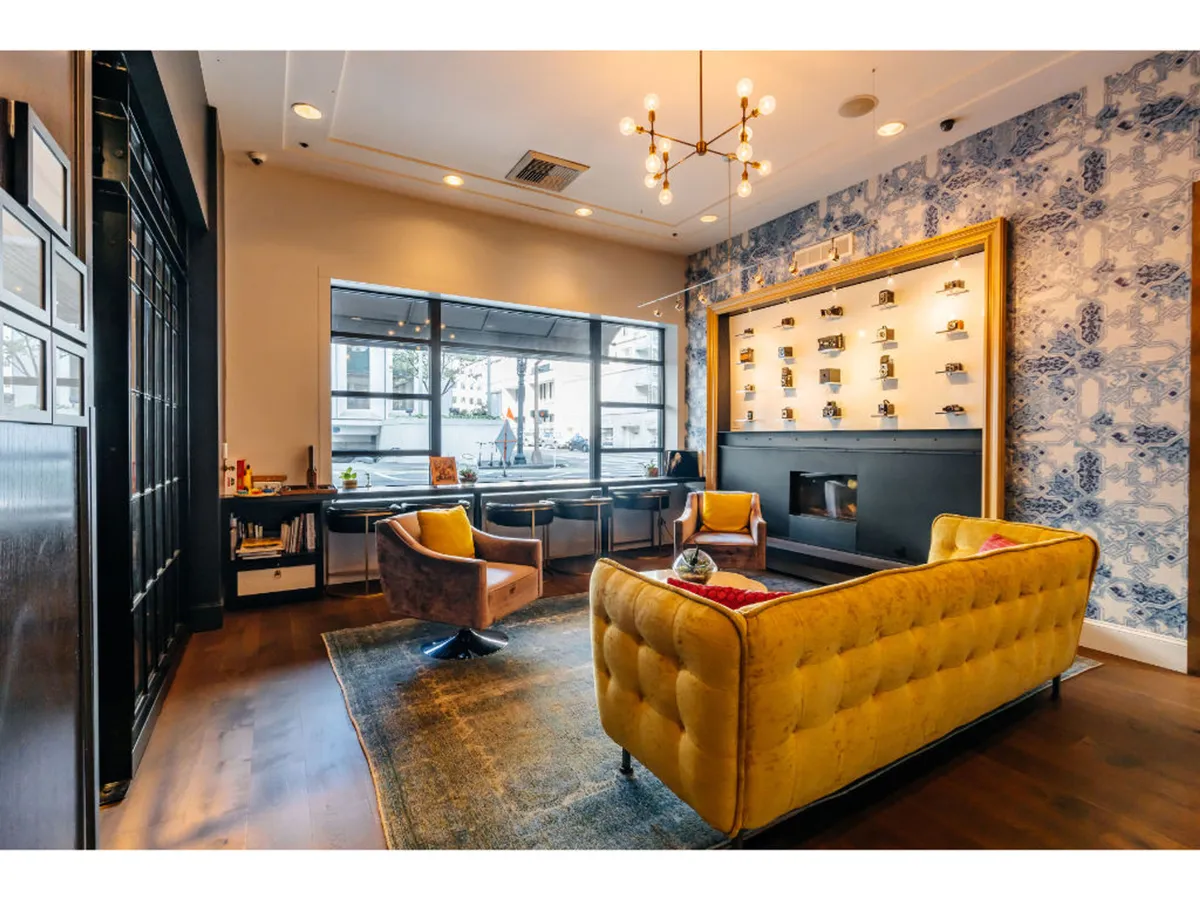 A stylish hotel lobby featuring a yellow tufted sofa set against blue patterned wallpaper, illuminated by a modern chandelier. A wooden table with chairs provides a welcoming environment for guests to meet, relax, or enjoy a coffee.