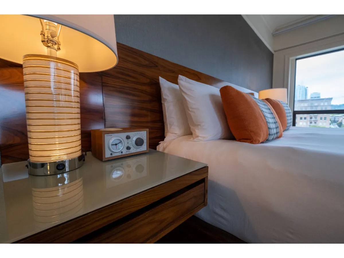 A tranquil scene focused on a comfortable bed with fluffy pillows, accompanied by a table lamp and an alarm clock on the nightstand. The overall atmosphere is restful, with soft lighting and simple, modern decor.