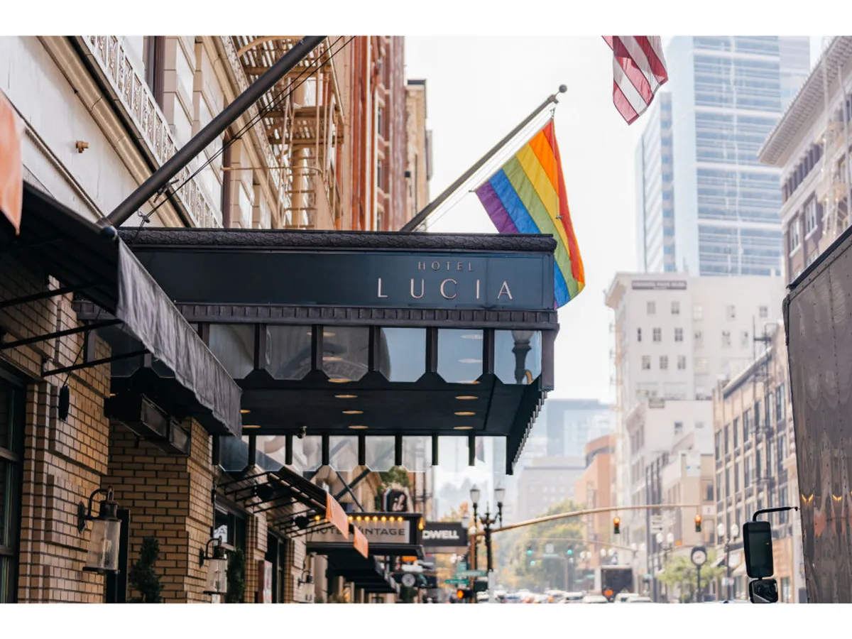 The exterior of Hotel Lucia adorned with a prominent hotel sign, rainbow flag, and American flag. Pedestrians are seen walking nearby, suggesting easy access to the city’s vibrant surroundings. The scene conveys an inviting and welcoming atmosphere at the hotel's entrance, with nearby cafes visible.