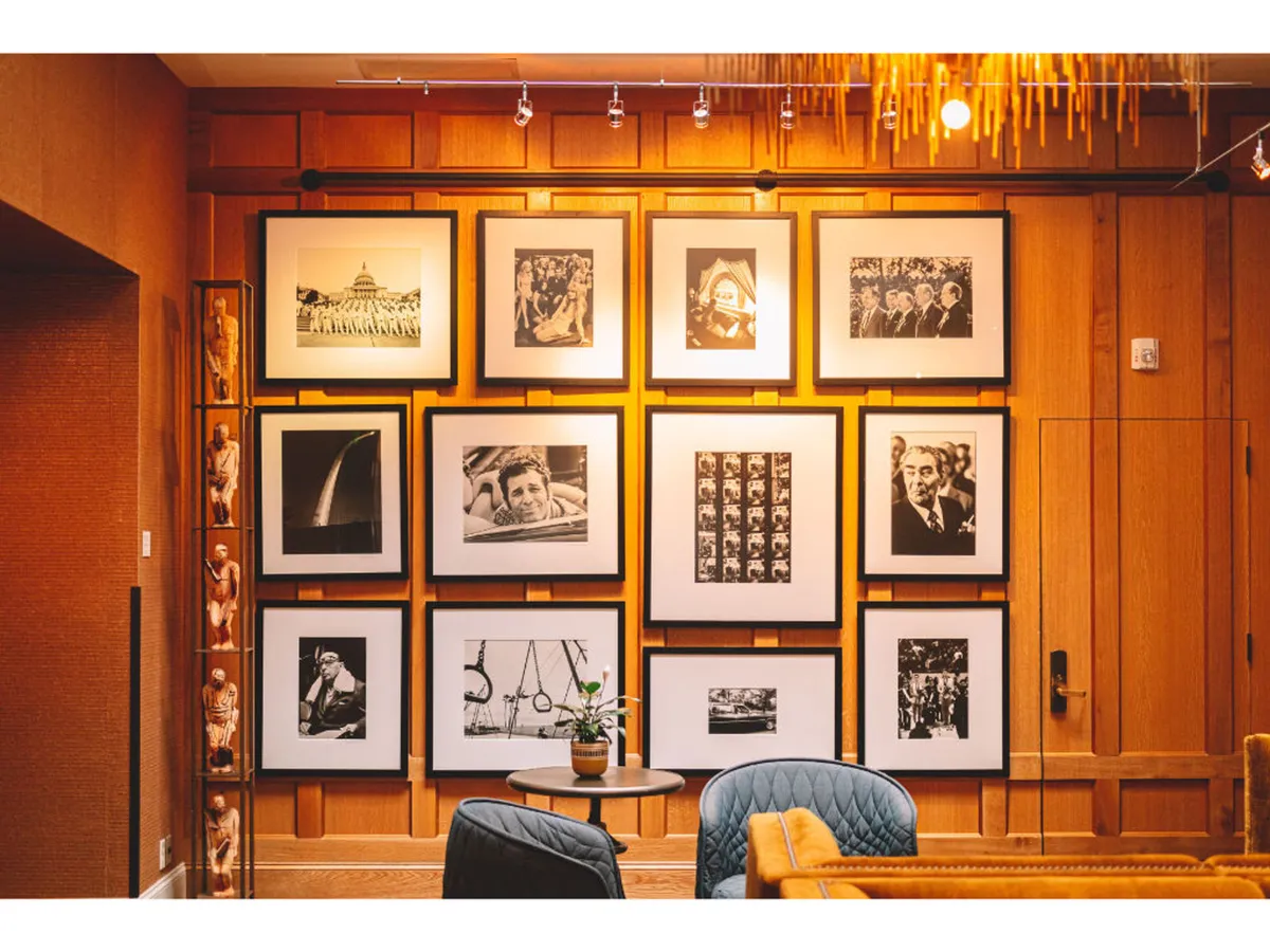 A cozy interior space featuring a gallery wall of framed photographs, blue upholstered chairs, a small round table, and warm wooden paneling. The setting invites visitors to relax, read, or engage in thoughtful conversation while surrounded by historical art.