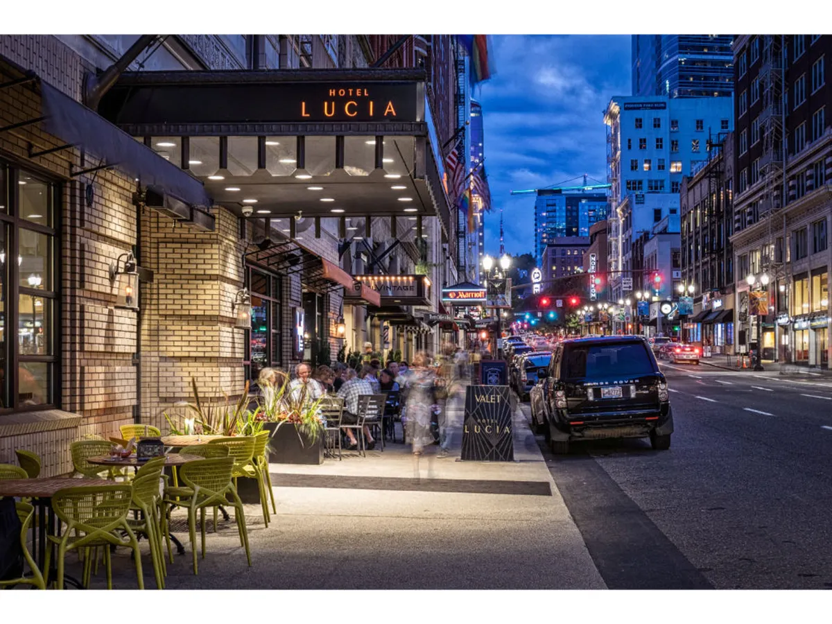 A view of the Hotel Lucia sign with an outdoor dining area and the city skyline in the background. People are seen dining and socializing, creating a sense of connection between the hotel and the vibrant downtown nightlife.