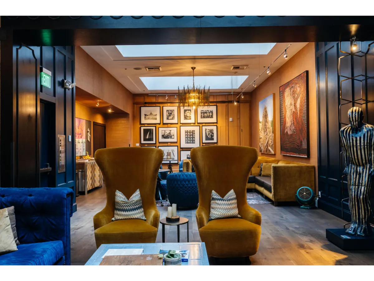 A vibrant lounge area with two large yellow armchairs, modern lighting, artwork on the walls, and a plush blue velvet couch. The space is bright and lively, ideal for guests to socialize, appreciate art, or participate in themed events.