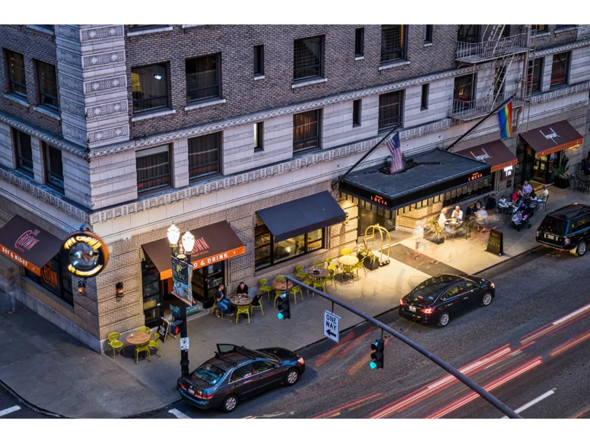 This image captures the inviting exterior of a café with outdoor seating and an illuminated patio area. Groups of people are seen dining outside, socializing, and enjoying evening drinks, while the surrounding cityscape and a nearby traffic light hint at the vibrant urban context.
