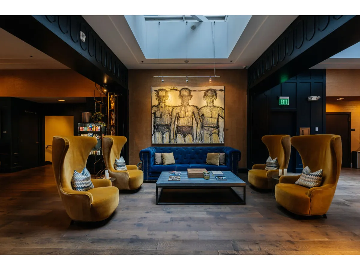 An inviting lounge area with a large blue sofa, yellow accent chairs, eye-catching artwork on the wall, and a coffee table. The atmosphere feels comfortable yet creative, encouraging both relaxation and appreciation of local art.