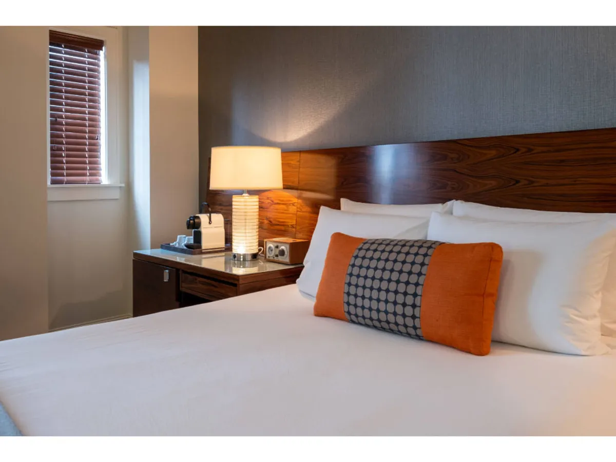 A serene guestroom featuring a plush queen bed adorned with decorative pillows, a nightstand topped with a modern lamp, and a window with blinds allowing natural light to filter in. The open arrangement offers clear floor space and an inviting, accessible atmosphere.