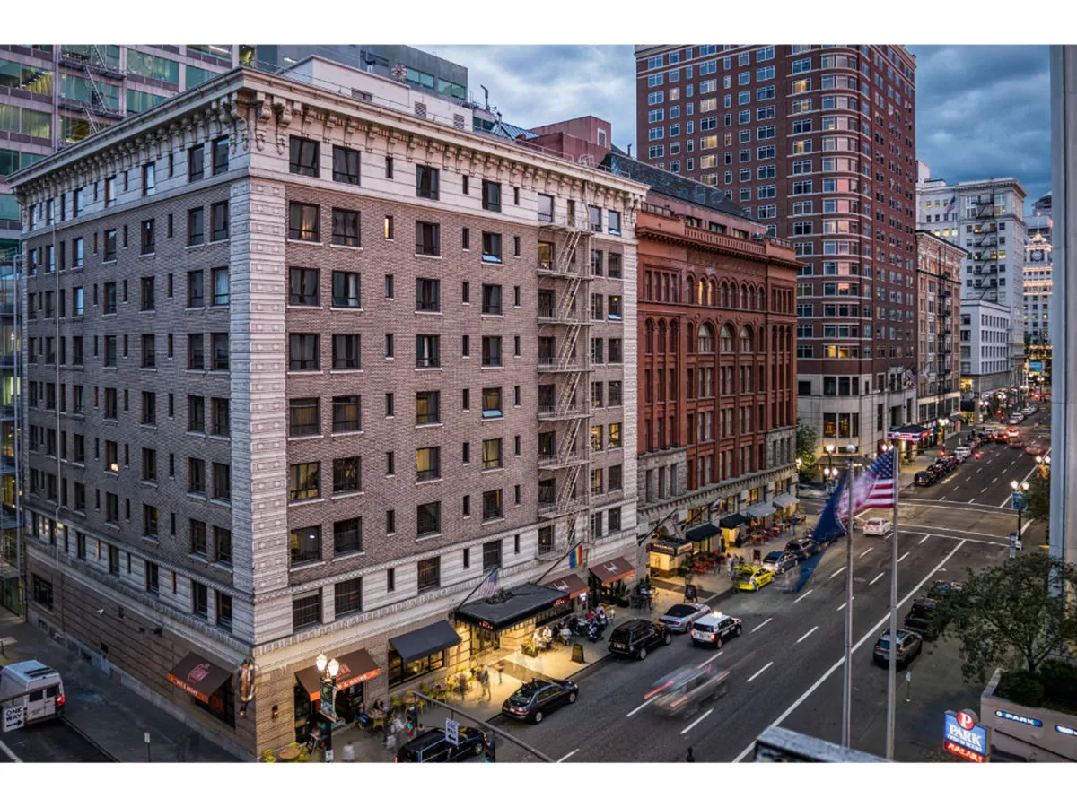 A bustling city street scene featuring a historic brick building, modern skyscrapers in the background, vibrant street cafes, and plenty of pedestrian activity. The energy of the neighborhood is palpable, reflecting the vibrancy and accessibility of the area.
